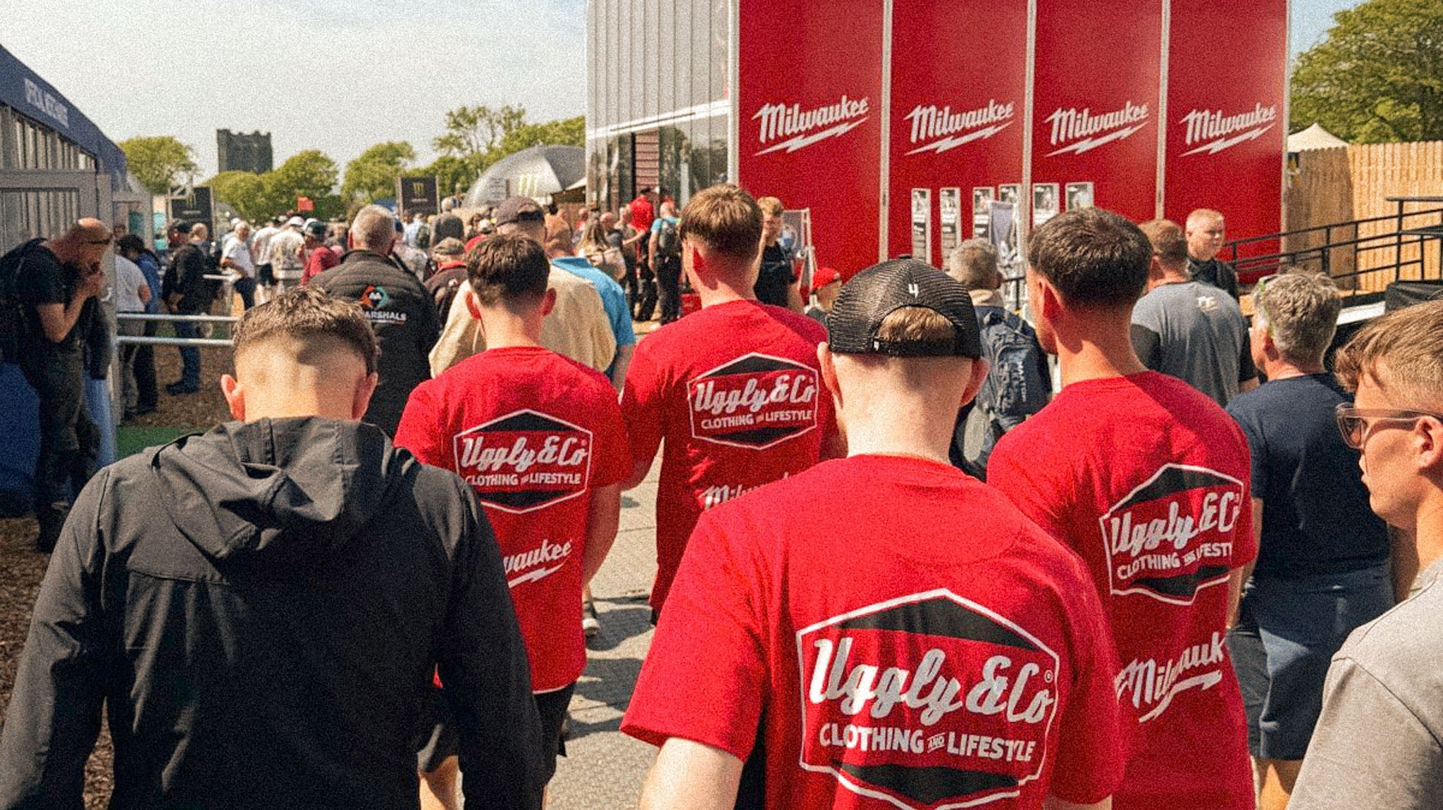 race fans wearing red Milwaukee collaboration t-shirts walking through a busy motorcycle event paddock on a sunny day