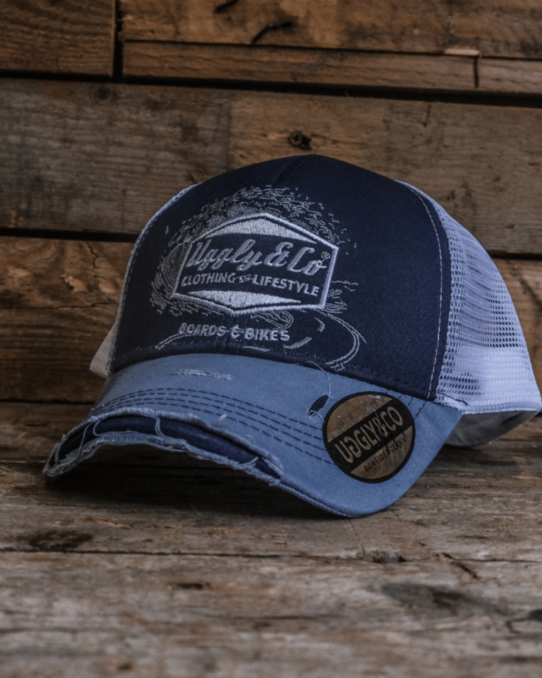 A blue trucker hat with a white print of the Uggly&Co diamond logo and a light blue wave texture on the front, featuring a navy blue front panel, a sun-faded light blue peak, and a breathable white mesh back, displayed on a wooden surface.