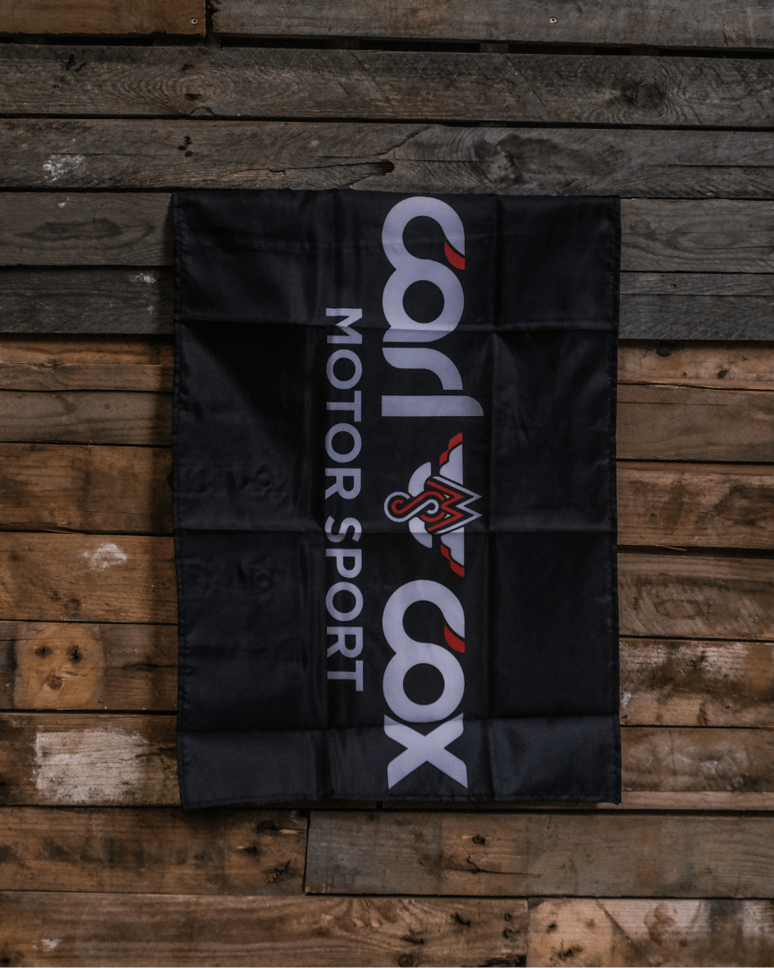 A rectangular black flag with the 'Carl Cox Motorsport' logo in white and red, displayed on a wooden surface.