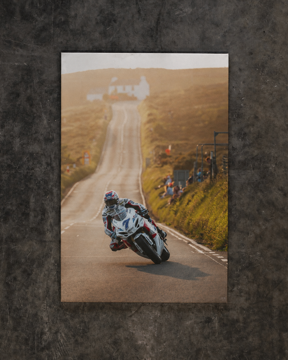 A vertical photographic print of a motorcyclist leaning into a turn on a winding road, displayed against a dark textured wall.