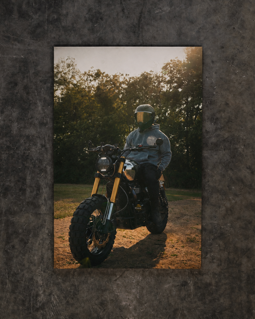 A vertical photographic print of a person in a helmet riding a motorcycle on a dirt path, surrounded by trees at sunset, displayed against a dark textured background.