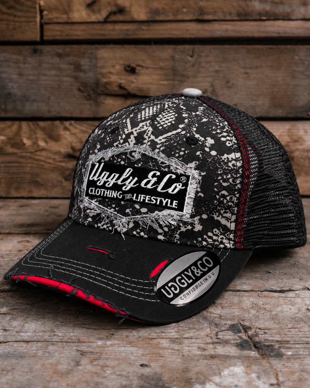 A black and white trucker hat with a distressed pattern on the front panel and brim, featuring a white 'Uggly & Co' logo patch. The back mesh is black with thin red stitching accents, and the hat is photographed on a rustic wooden plank surface.