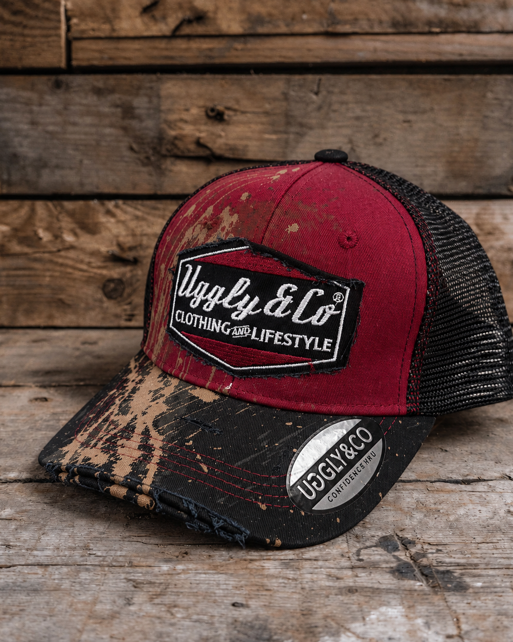 A distressed trucker hat with a red front panel, black mesh back, and a black brim featuring a brown and beige Mud splatter motorcross pattern. The front has a black patch with the text 'Uggly & Co CLOTHING AND LIFESTYLE'.