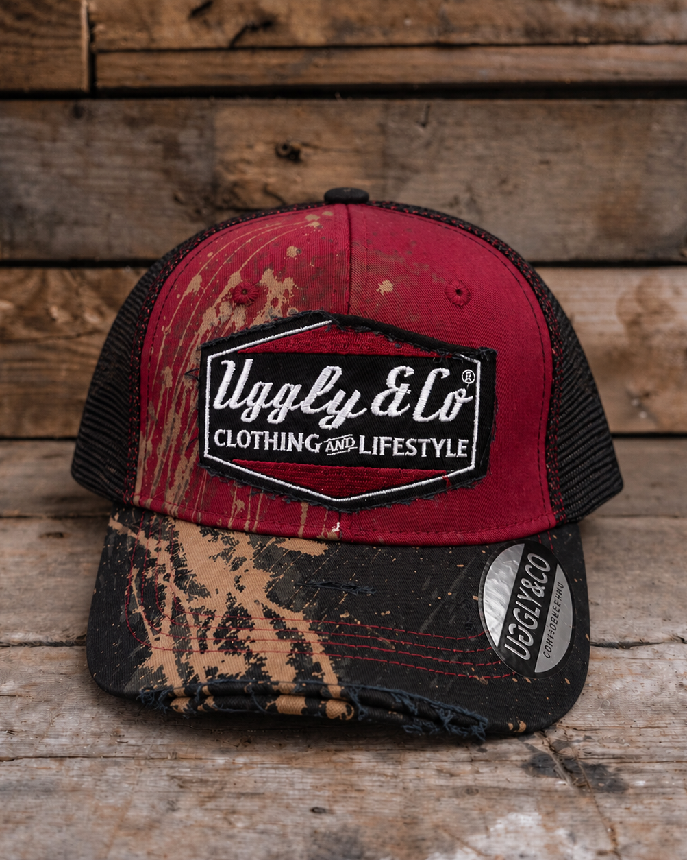 A trucker-style cap with a distressed red front panel, black mesh back, and a camouflage-patterned brim, displayed on a rustic wooden surface.