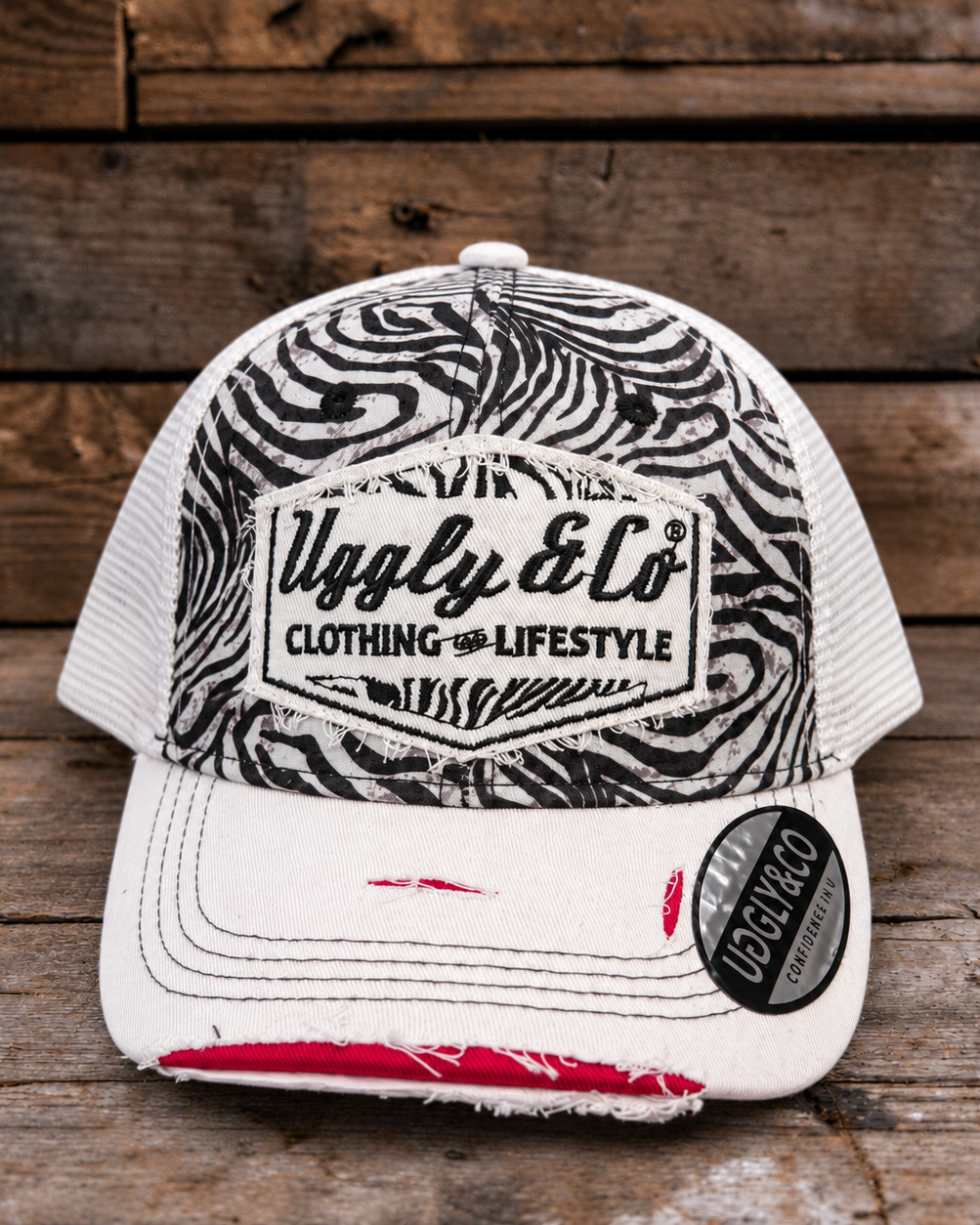 A distressed trucker cap with a black and white zebra print front panel, white mesh back, and a white brim with frayed detailing and a red accent on the underside. The front features a patch for 'Uggly & Co Clothing & Lifestyle' and a circular 'UGGLY & CO' sticker on the brim, set against a rustic wooden plank background.