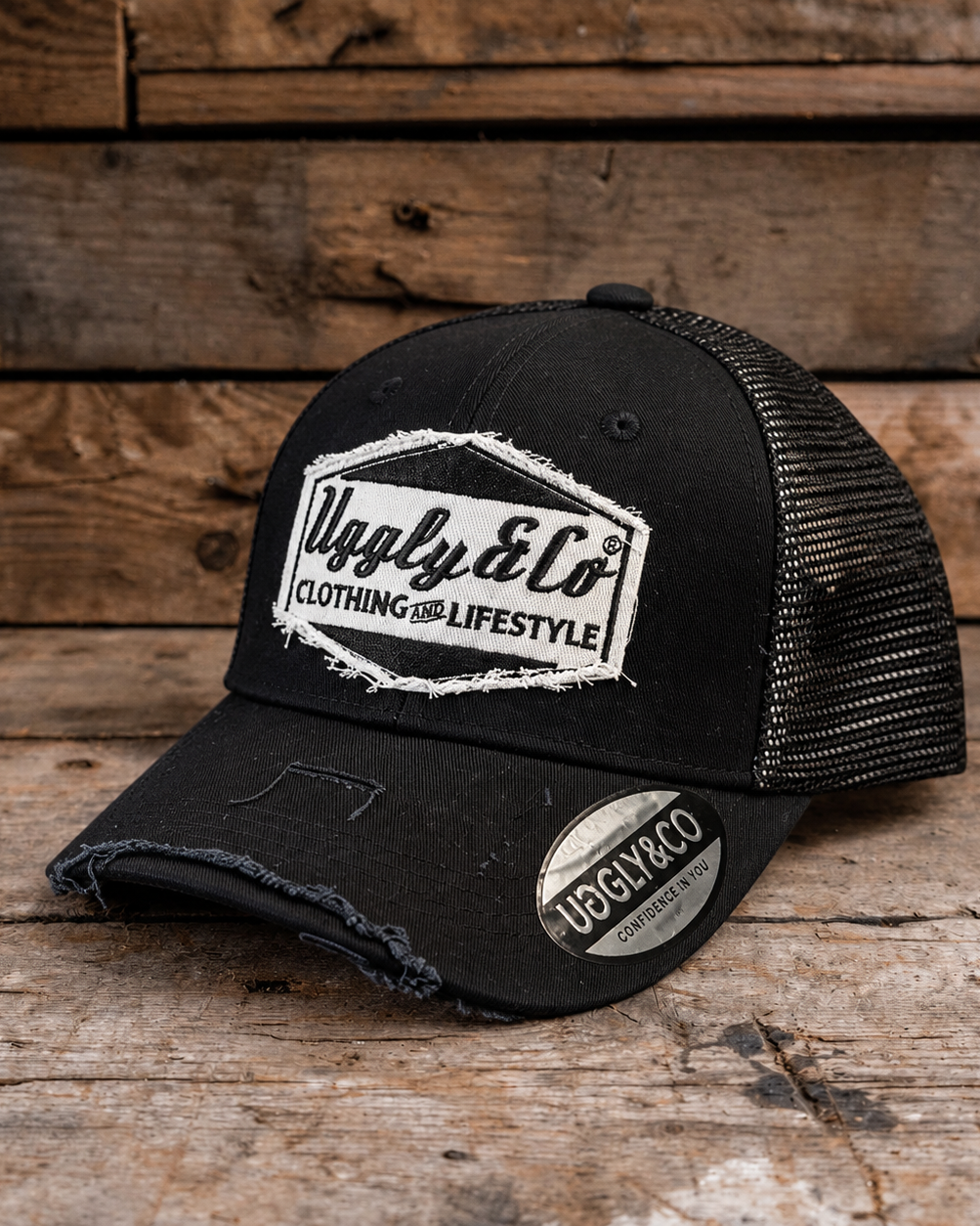 A black trucker-style cap with distressed detailing, a white embroidered 'Uggly & Co' logo on the front, and a matching sticker on the brim, displayed on a rustic wooden surface.