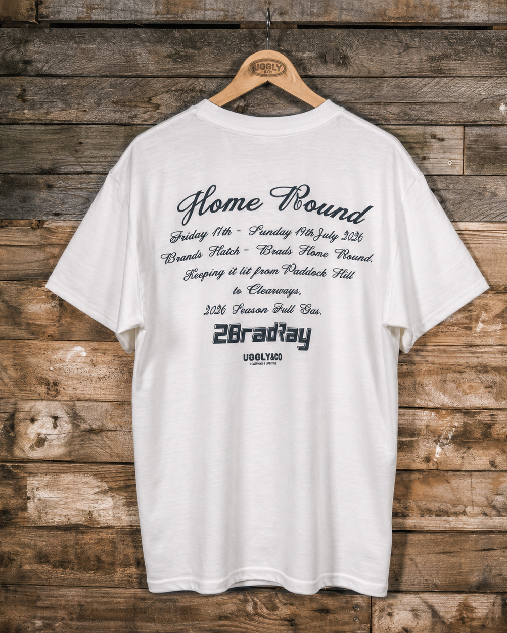 A white short-sleeve T-shirt hanging on a wooden hanger against a rustic brown wood plank wall. The back of the shirt features black printed text celebrating a horse race victory, including 'Home Round', dates, track details, and '2BradRay USOLYED'.
