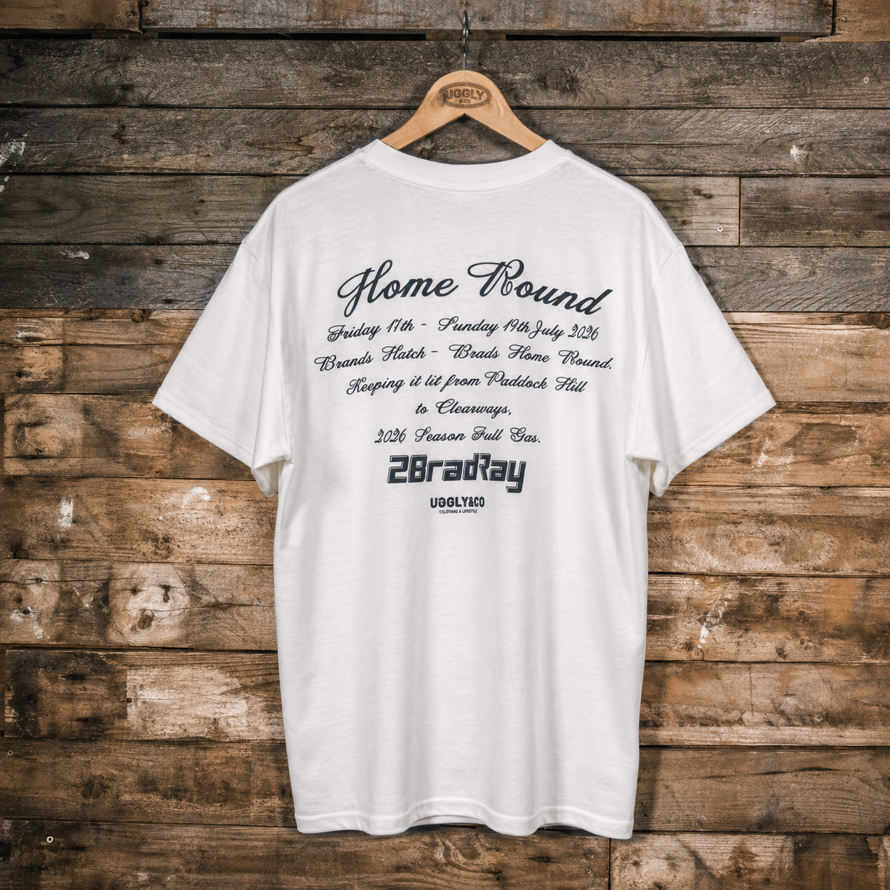 A white short-sleeve T-shirt hanging on a wooden hanger against a rustic brown wood plank wall. The back of the shirt features black printed text celebrating a horse race victory, including 'Home Round', dates, track details, and '2BradRay USOLYED'.