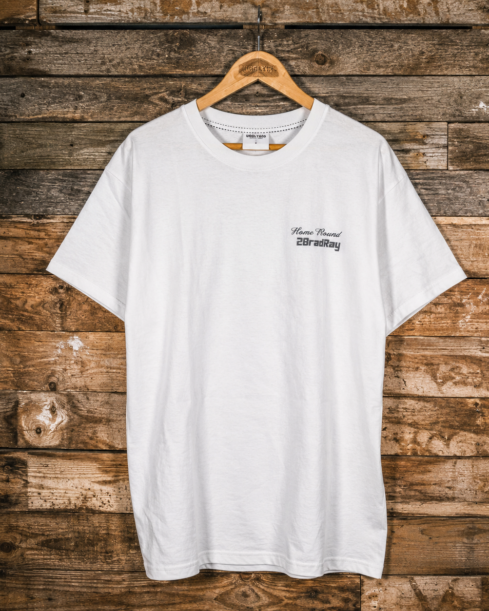 A white short-sleeve crewneck t-shirt with a small black graphic on the left chest, hanging on a wooden hanger against a rustic brown wood plank wall.