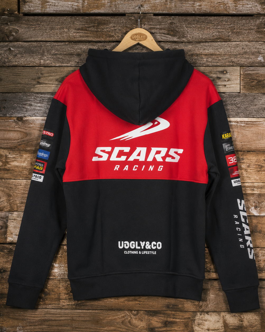 A black and red hooded sweatshirt shown from the back, hanging on a wooden hanger against a rustic wood plank wall. The hoodie features a bold red upper back with a large white 'SCARS RACING' logo, black sleeves, and multiple sponsor-style patches on the sleeves.