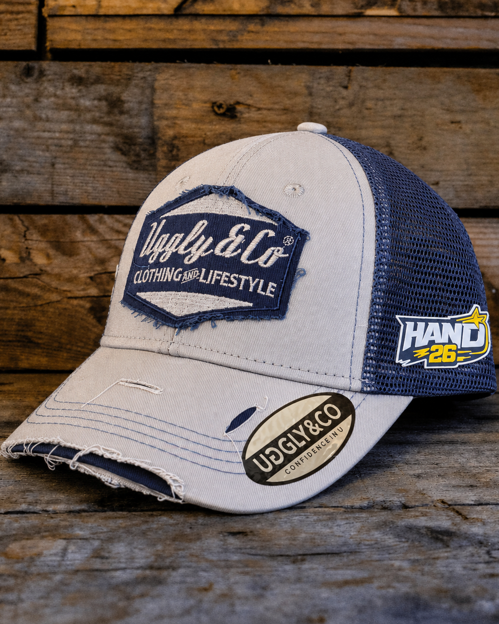 A distressed trucker cap with a light beige front and brim, navy blue mesh back panels, and embroidered patches including 'Ugly & Co' and 'HAND 26', displayed on a rustic wooden surface.