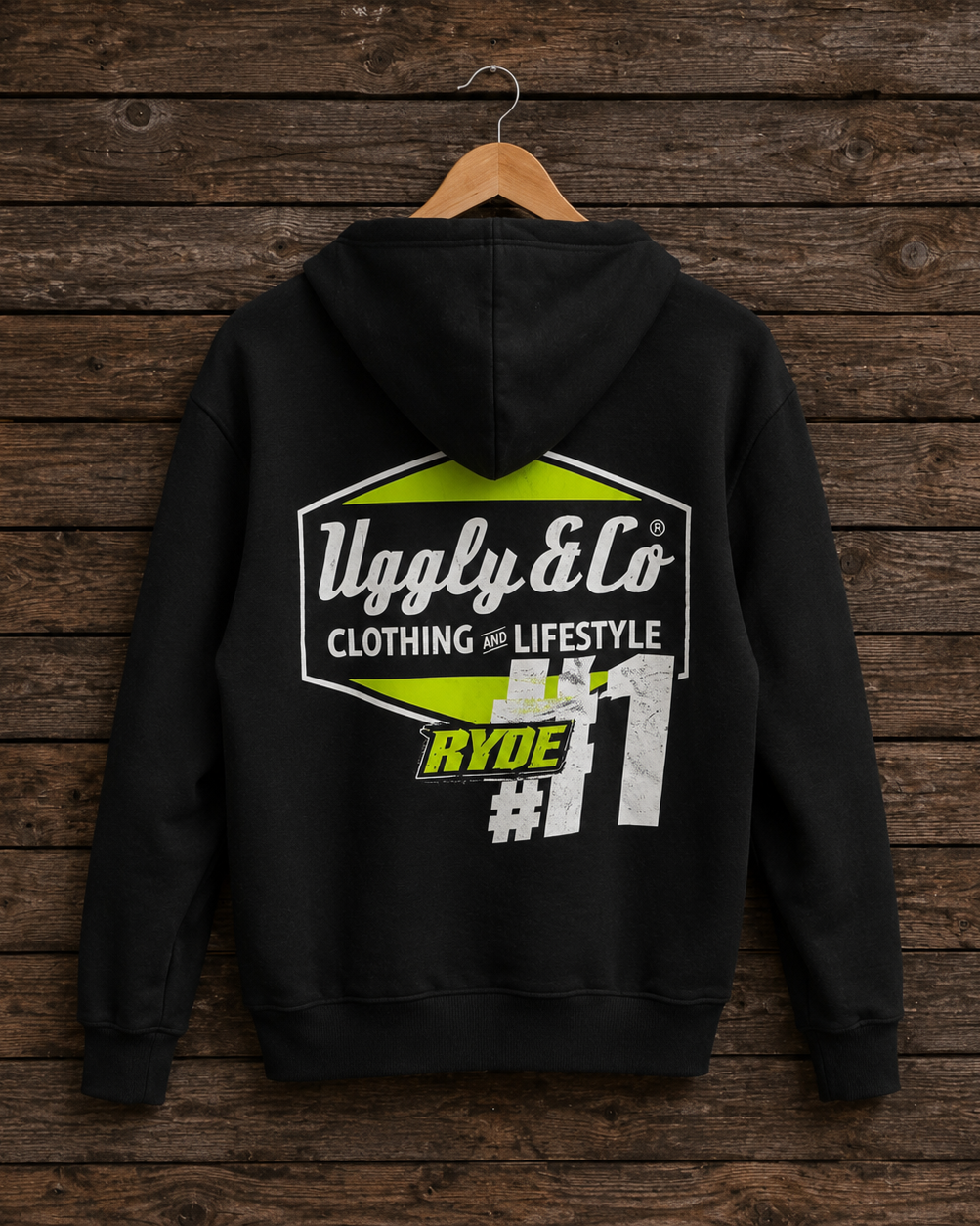 A black hoodie hanging on a wooden hanger against a dark rustic wood plank wall. The back of the hoodie features a large graphic print with the text 'Ugly & Co CLOTHING LIFESTYLE', a lime green hexagon shape, and 'RYDE #11'.