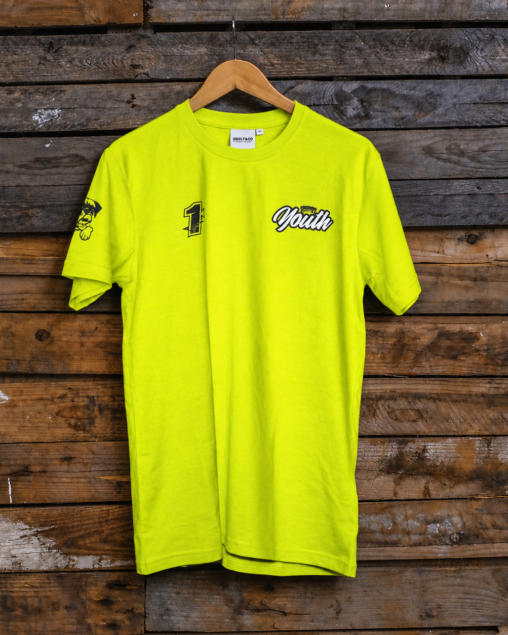 A bright neon yellow t-shirt hanging on a wooden hanger against a rustic dark wood plank wall. The shirt features a black graphic '1' on the left chest, a small mascot logo on the right sleeve, and 'Youth' in white script with a black outline across the center chest.