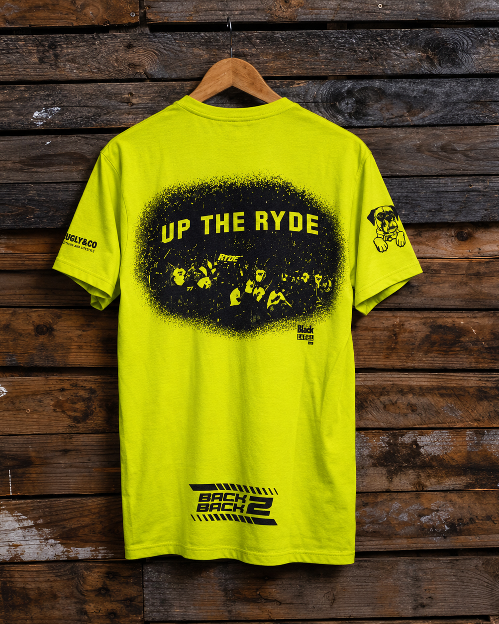 A bright neon yellow t-shirt hanging on a wooden hanger against a rustic dark wood plank wall. The back of the shirt features a large black graphic with yellow text reading 'UP THE RYDE' inside a distressed black circle, a small black graphic on the right sleeve, and a black graphic near the bottom hem.
