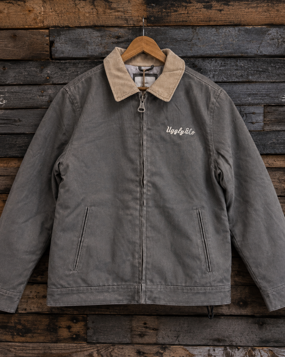 A gray canvas utility jacket with a tan corduroy collar, hanging on a wooden hanger against a rustic dark wood plank wall. The jacket features a front zipper, zippered side pockets, and white embroidered script on the left chest.