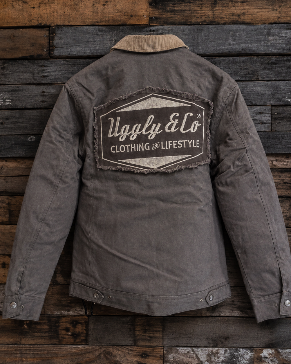 A dark gray utility jacket with a tan collar, shown from the back against a rustic wooden plank wall. The back features a large distressed patch with the brand name 'Uggly & Co Clothing and Lifestyle'.