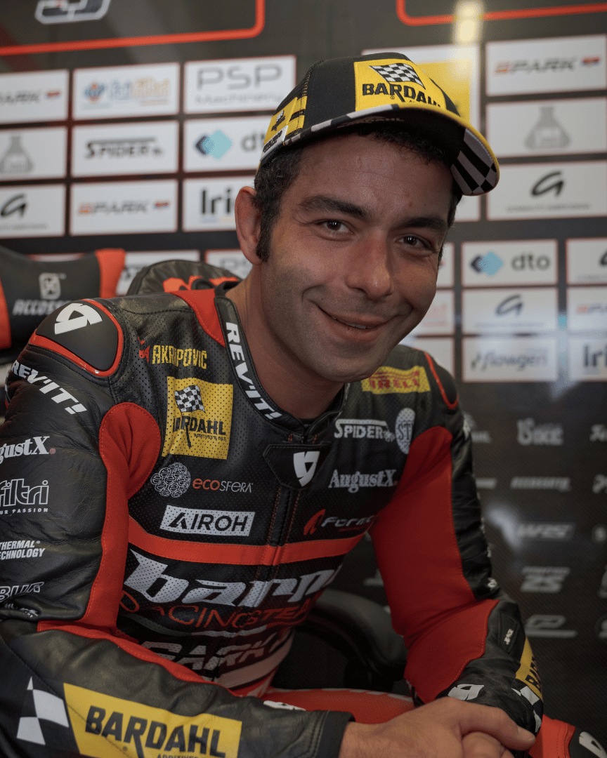 Danilo Pertrucci smiling at the camera while wearing a yellow and black Uggly&Co Bardahl Danilo Pertrucci #9 merchandise trucker hat