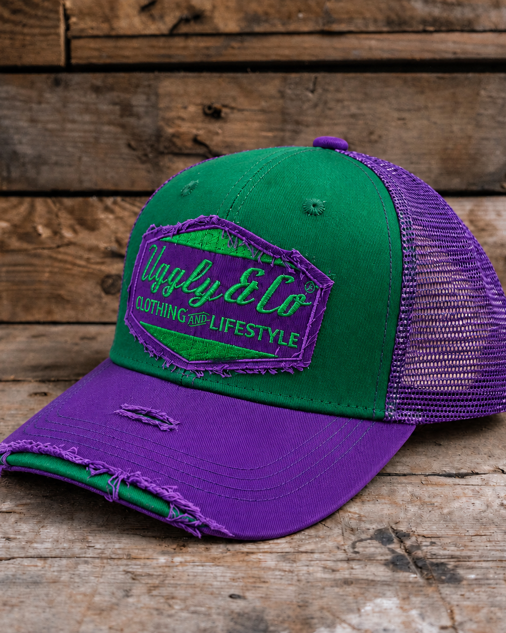 A two-tone green and purple trucker cap with a distressed brim, resting on a rustic wooden surface against a wooden plank background. The front panel features a green and purple embroidered patch reading 'Uggly & Co CLOTHING FOR LIFESTYLE'.