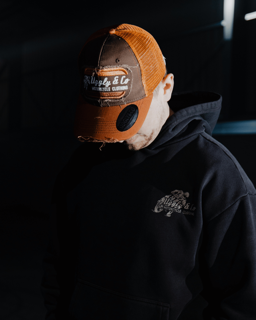 A model looking down while wearing a brown trucker hat with an orange brim, featuring a flat track Uggly&Co logo patch on the front at night