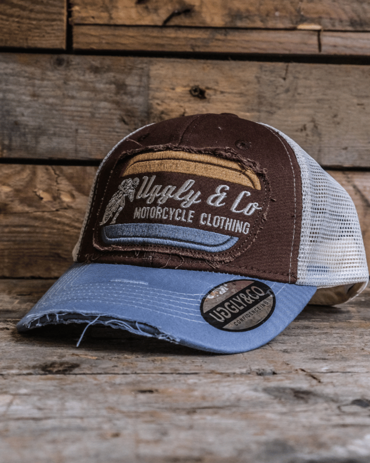 A dusty blue trucker hat with a brown front panel, featuring a vintage flat track Uggly&Co logo, placed on a wooden surface.