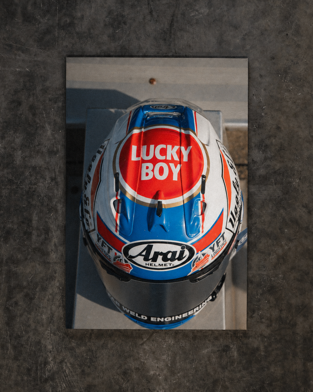 A top-down view of a racing helmet with a reflective mirror finish, featuring prominent 'Lucky Boy' and 'Arai' branding, set against a dark textured concrete background.