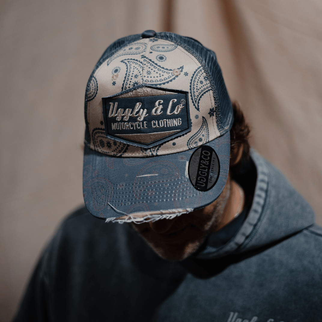A model in a hoodie looking down while wearing a blue and beige Bandana Diamond Trucker Hat with a motorcycle retro bandana print and the Uggly&Co logo on the front, with a blurred background