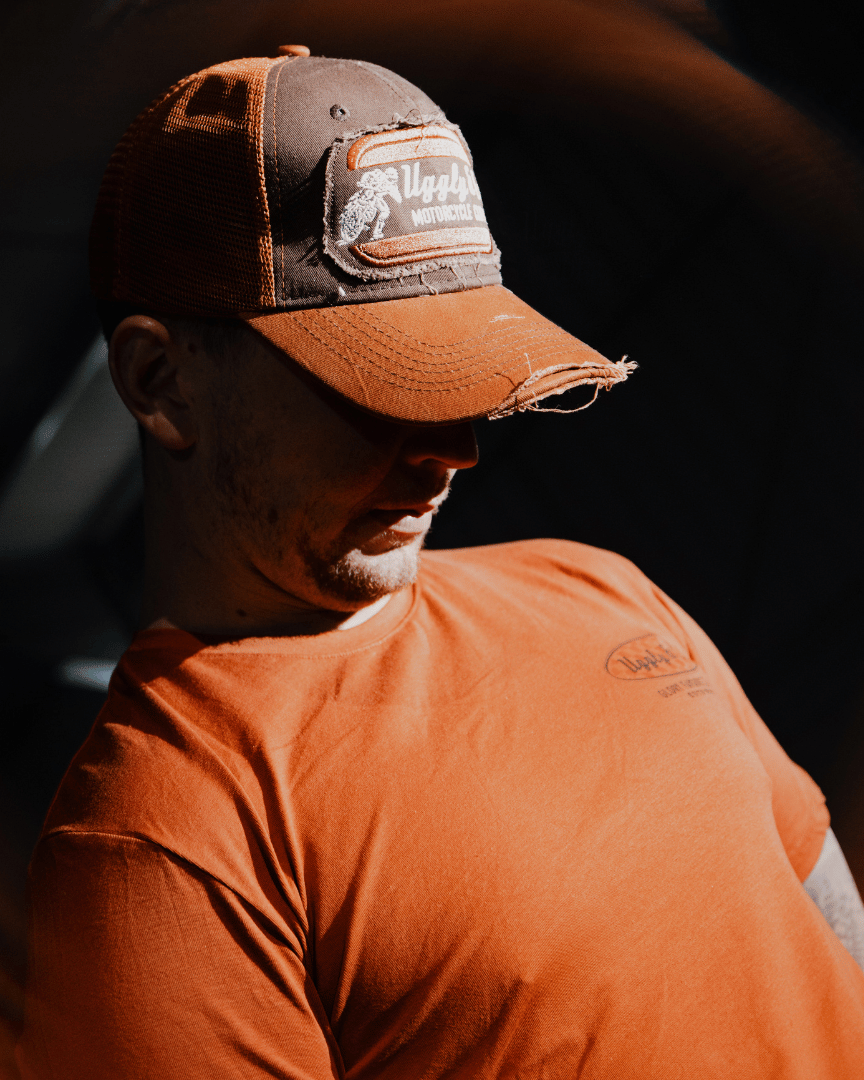 A model sitting down while wearing a brown trucker hat with an orange brim, featuring a flat track Uggly&Co logo patch on the front at night