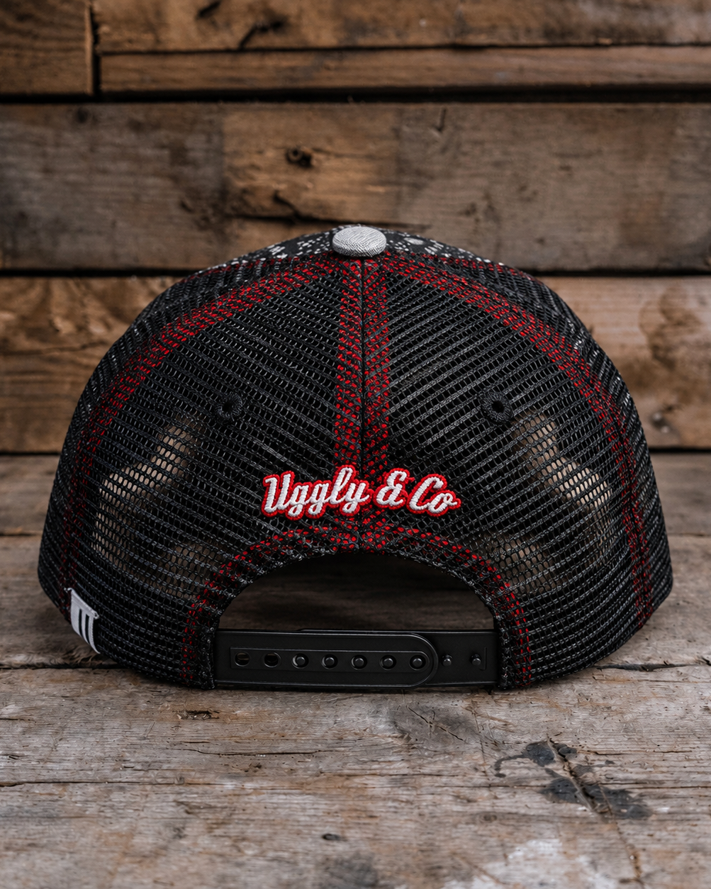A black trucker-style cap with red mesh trim and red embroidered 'Uggly & Co' text on the back, photographed on a rustic wooden surface with a matching wood plank background.