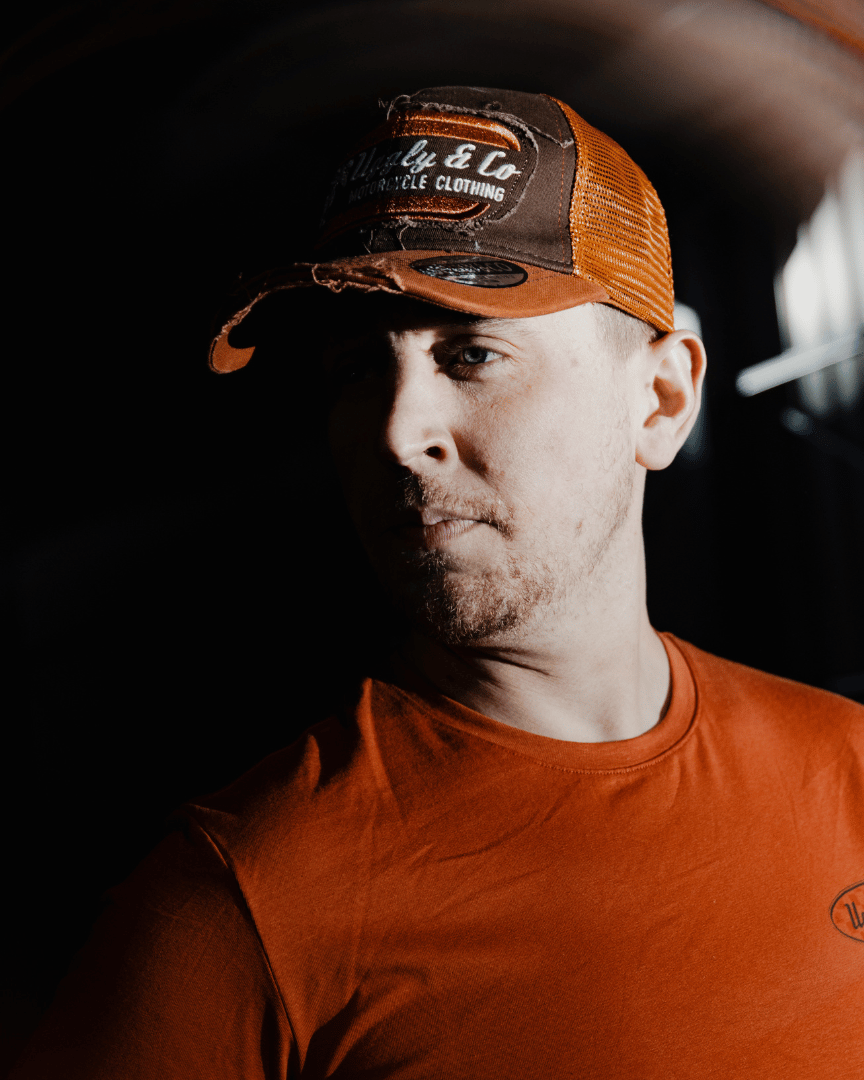 A model looking left while wearing a brown trucker hat with an orange brim, featuring a flat track Uggly&Co logo patch on the front at night