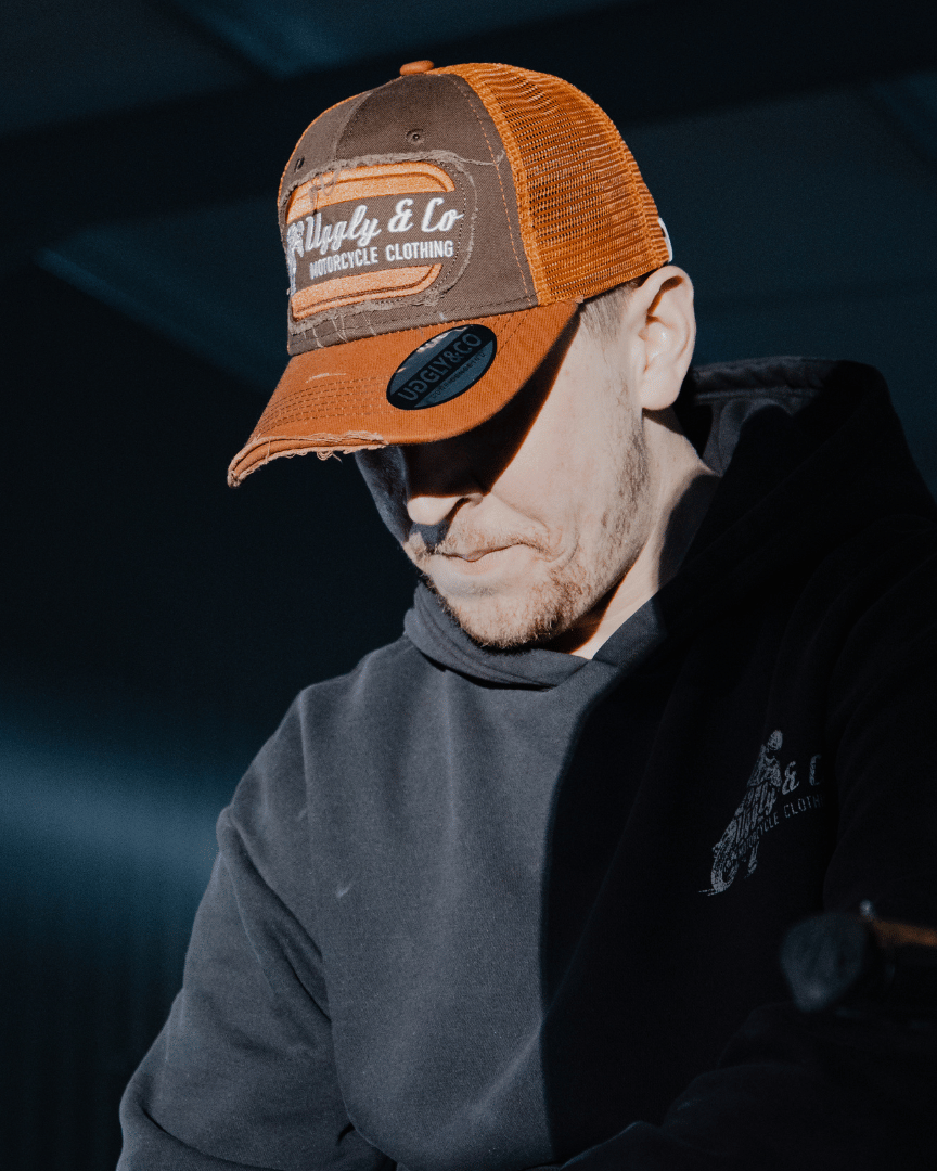 A model wearing a brown trucker hat with an orange brim, featuring a flat track Uggly&Co logo patch on the front at night
