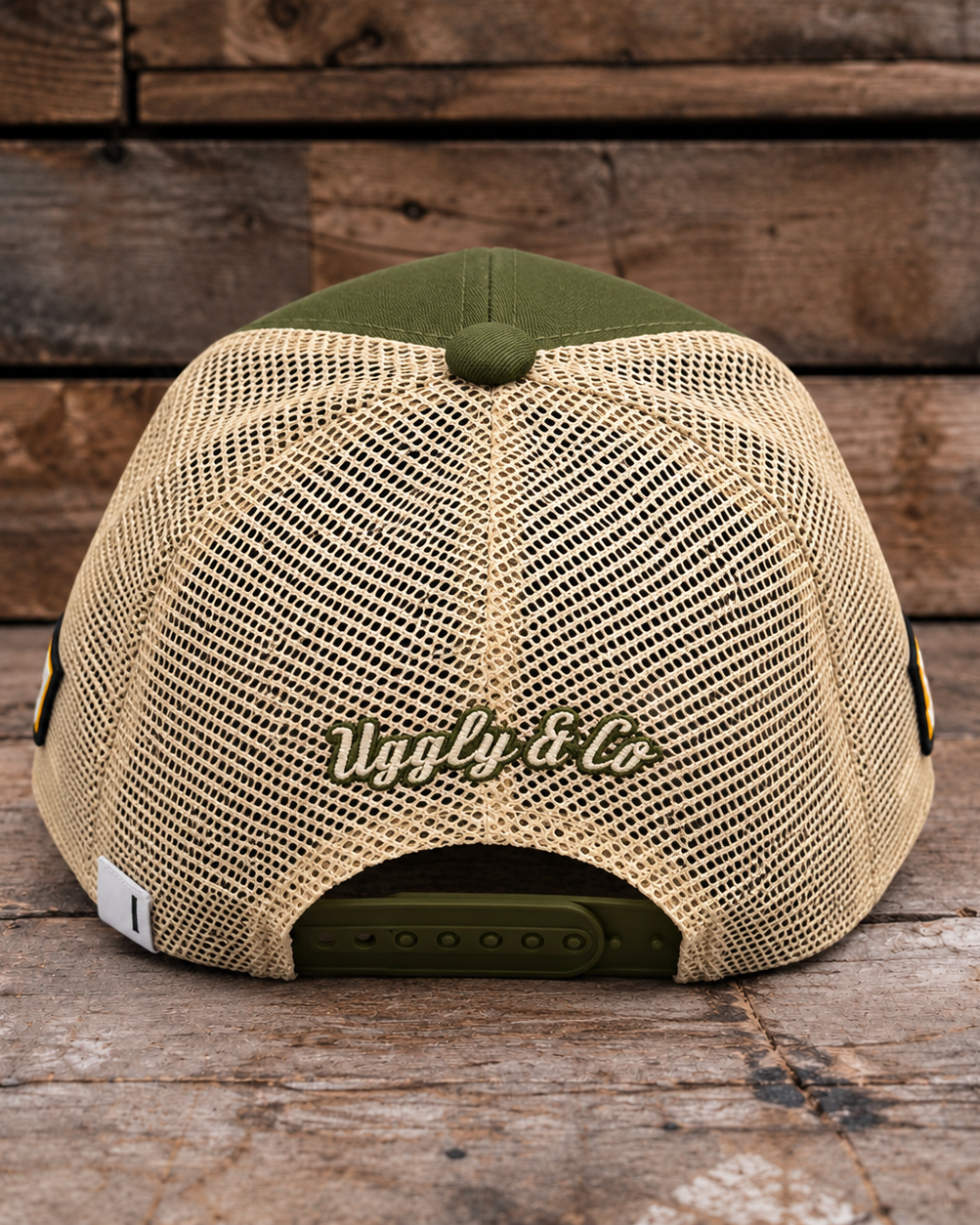 A beige mesh-back trucker cap with an olive green front panel, button, and adjustable strap, featuring 'Uggly & Co' embroidered on the back, set against a rustic wooden plank background.