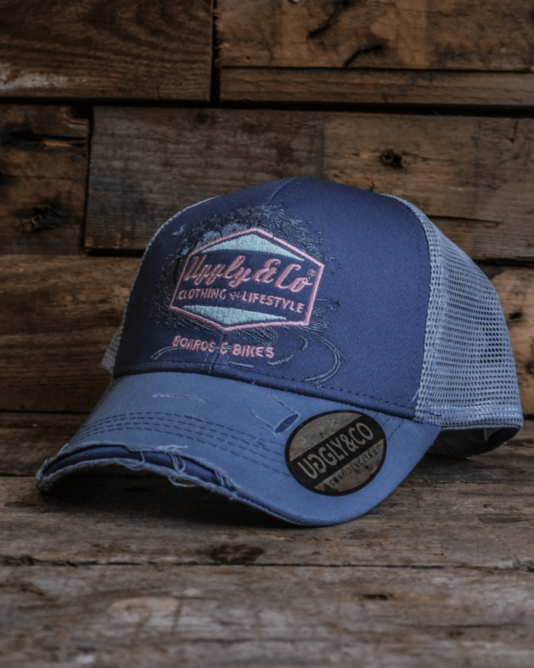A trucker hat with a purple front panel and a blue mesh back, featuring a pink diamond Uggly&Co logo and the text 'Boards & Bikes' with a wooden background
