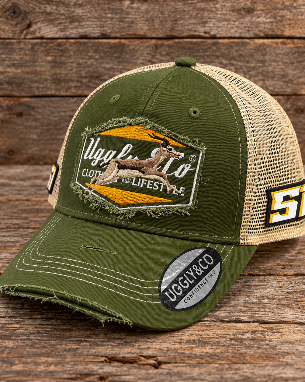An olive green and beige trucker hat with a distressed front patch reading 'Uggly Co' and a springbok South Africa graphic, displayed on a rustic wooden surface.