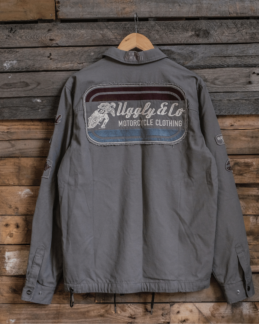 A gray motorcycle outerwear jacket with Flattrack and Uggly&Co branding, featuring a dual zip and button-up closure, displayed on a wooden background.