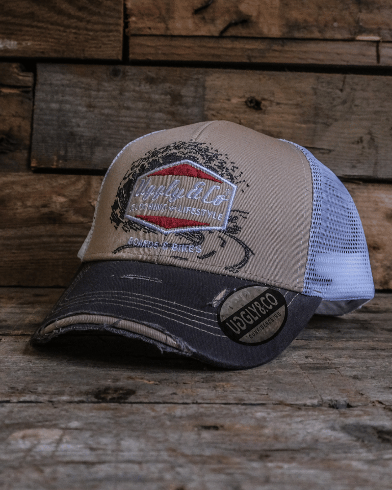 A beige and brown trucker cap with a diamond Uggly&Co logo and wave texture design, featuring a white mesh back and a curved peak, displayed against a wooden background.
