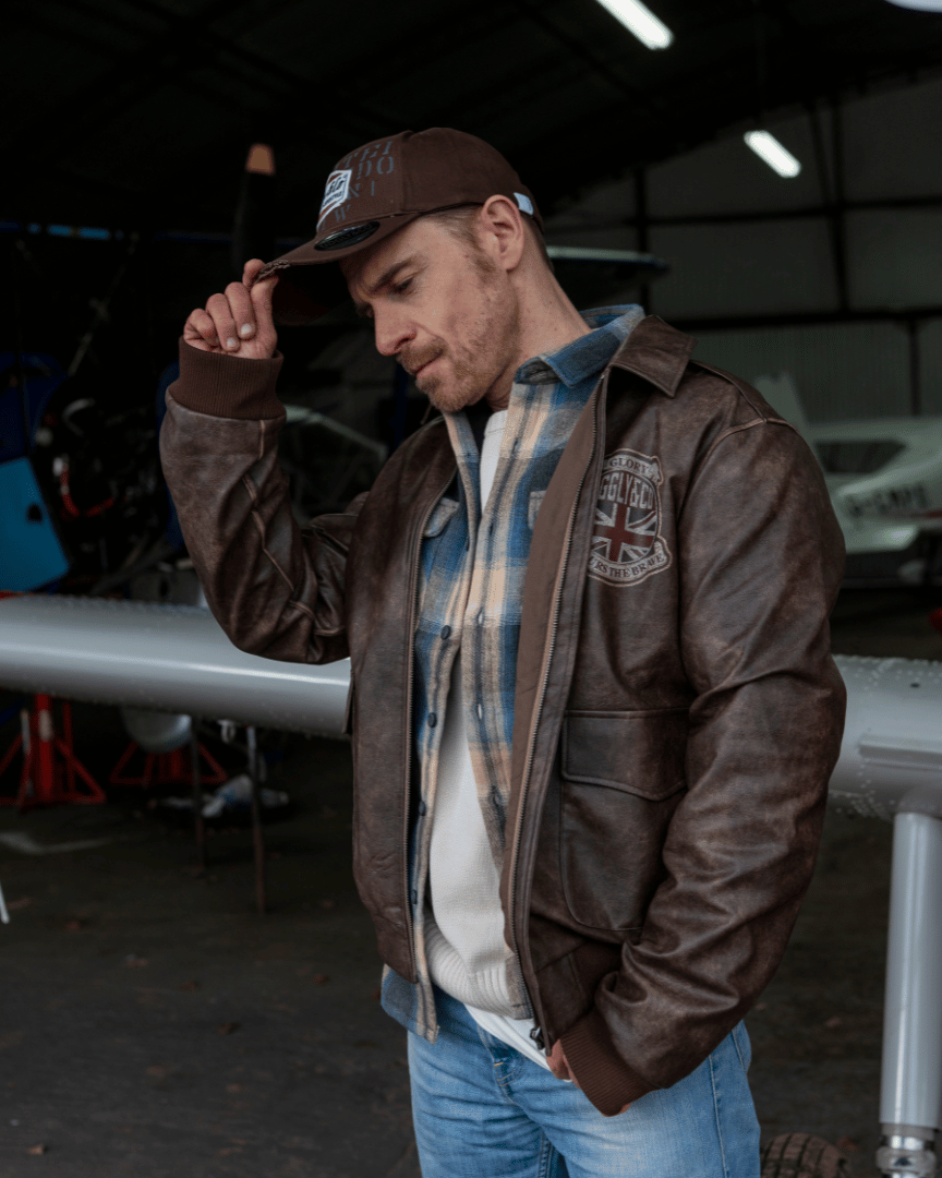 A man holding the peak of a brown trucker hat with 'Uggly & Co' branding with a plane in the background