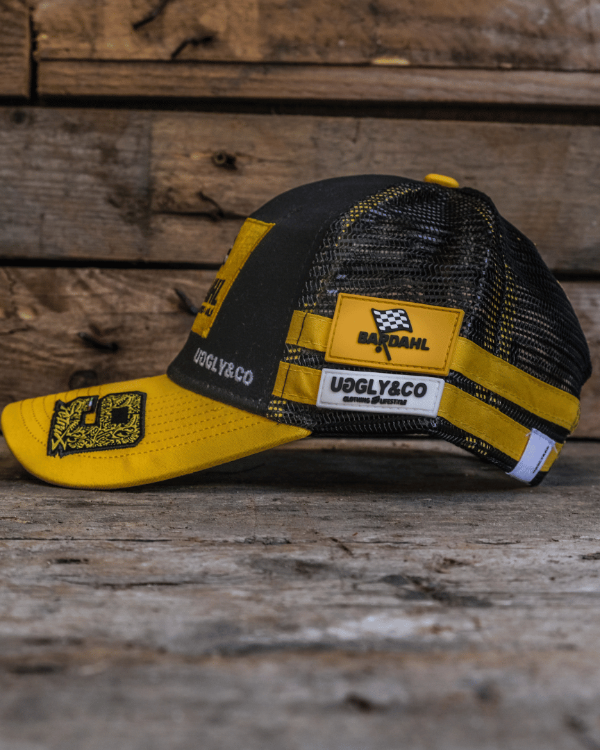 Side view of a yellow and black Uggly&Co Bardahl Danilo Pertrucci #9 merchandise trucker hat on a wooden surface