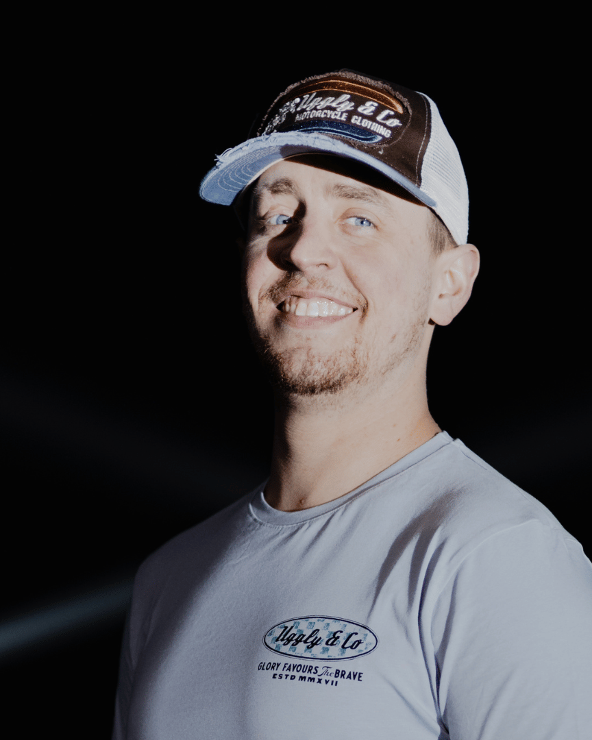 A model smiling at the camera while wearing a dusty blue trucker hat with a brown front panel, featuring a vintage flat track Uggly&Co logo