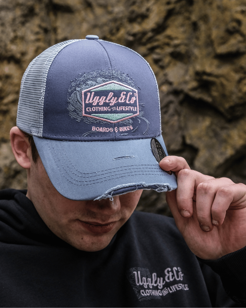A model looking down while wearing a trucker hat with a purple front panel and a blue mesh back, featuring a pink diamond Uggly&Co logo and the text 'Boards & Bikes' with a blurred rocky background
