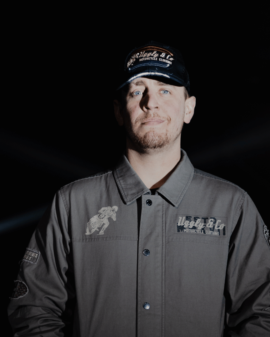 Front view of a model wearing an all-black trucker hat with a vintage-inspired flat track racing logo in yellow and blue on the front at night