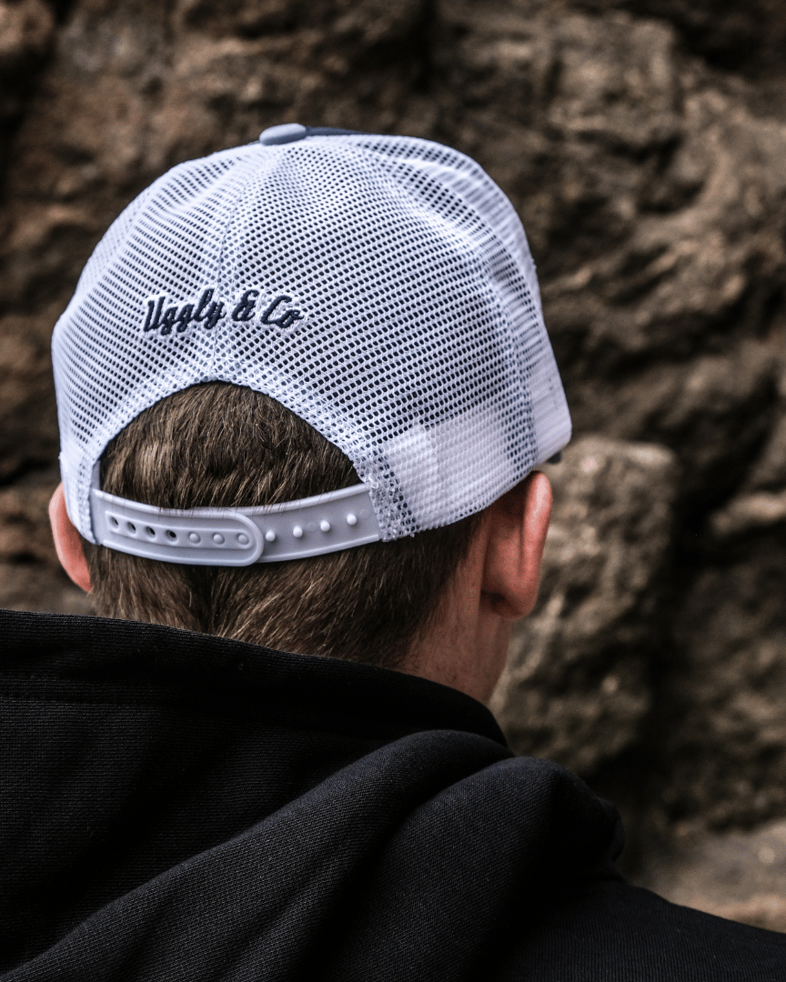 A rear view of a model wearing a blue trucker hat with a white print of the Uggly&Co diamond logo and a light blue wave texture on the front, featuring a navy blue front panel, a sun-faded light blue peak, and a breathable white mesh back
