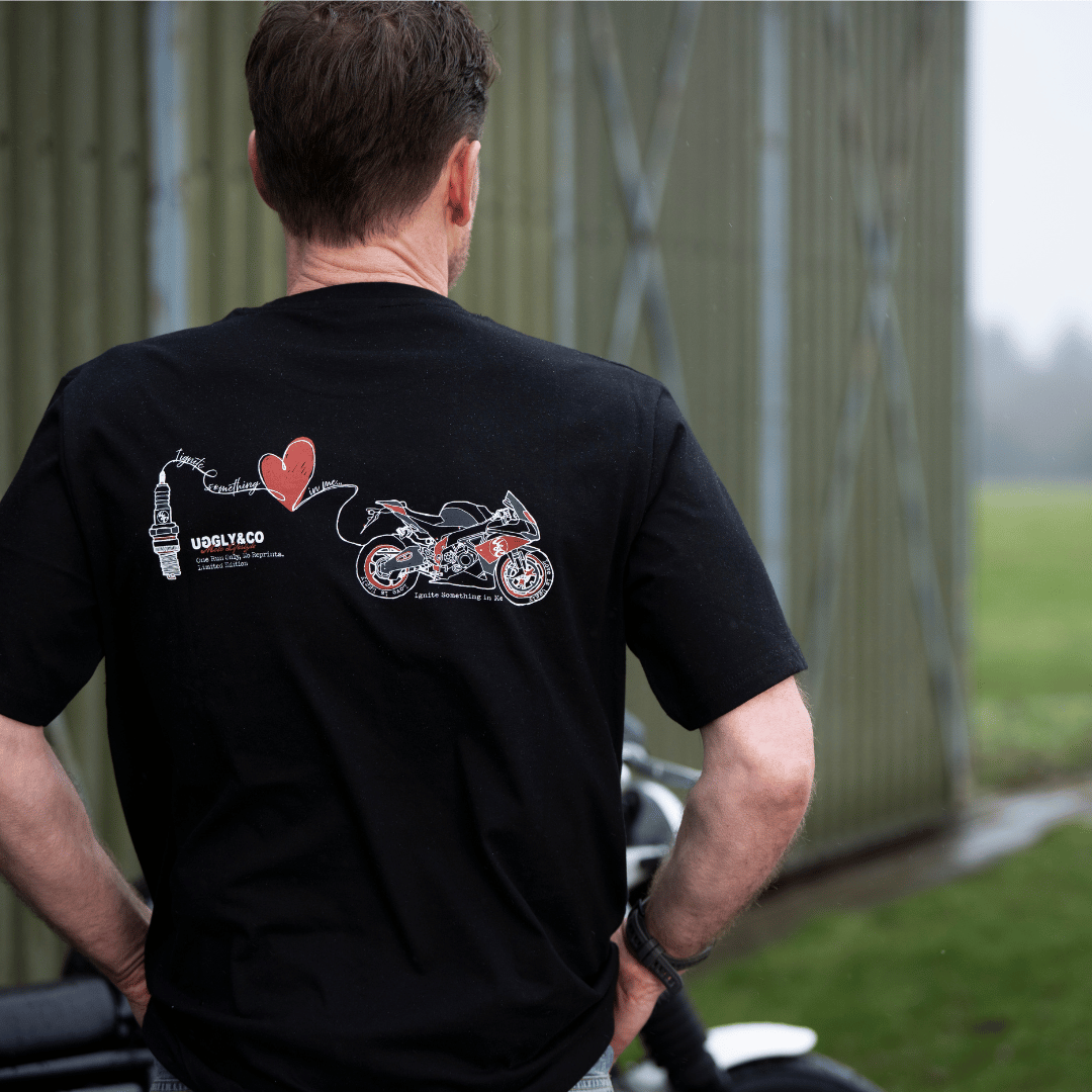Rear view of a model wearing a black t-shirt with a motorcycle, love heart and spark plug graphic with a background blurred