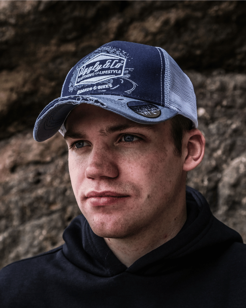 A front view of a model wearing a blue trucker hat with a white print of the Uggly&Co diamond logo and a light blue wave texture on the front, featuring a navy blue front panel, a sun-faded light blue peak, and a breathable white mesh back.