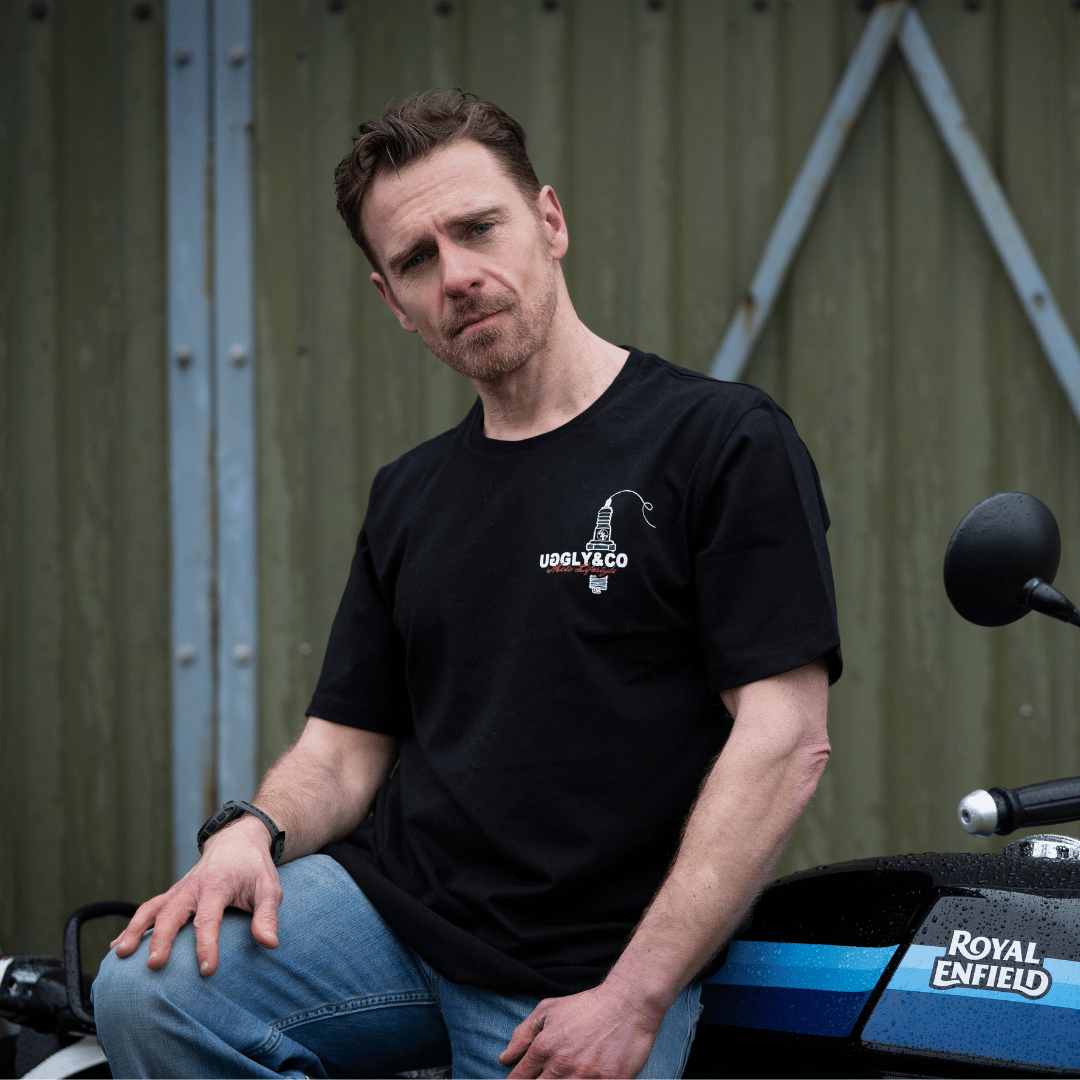 Front view of a model wearing a black t-shirt with a spark plug graphic and the Uggly&Co logo while sitting on a motorcycle and looking at the camera