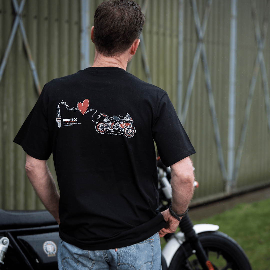 Rear view of a model wearing a black t-shirt with a spark plug, heart and motorcycle graphic and the Uggly&Co logo with a motorcycle in the background