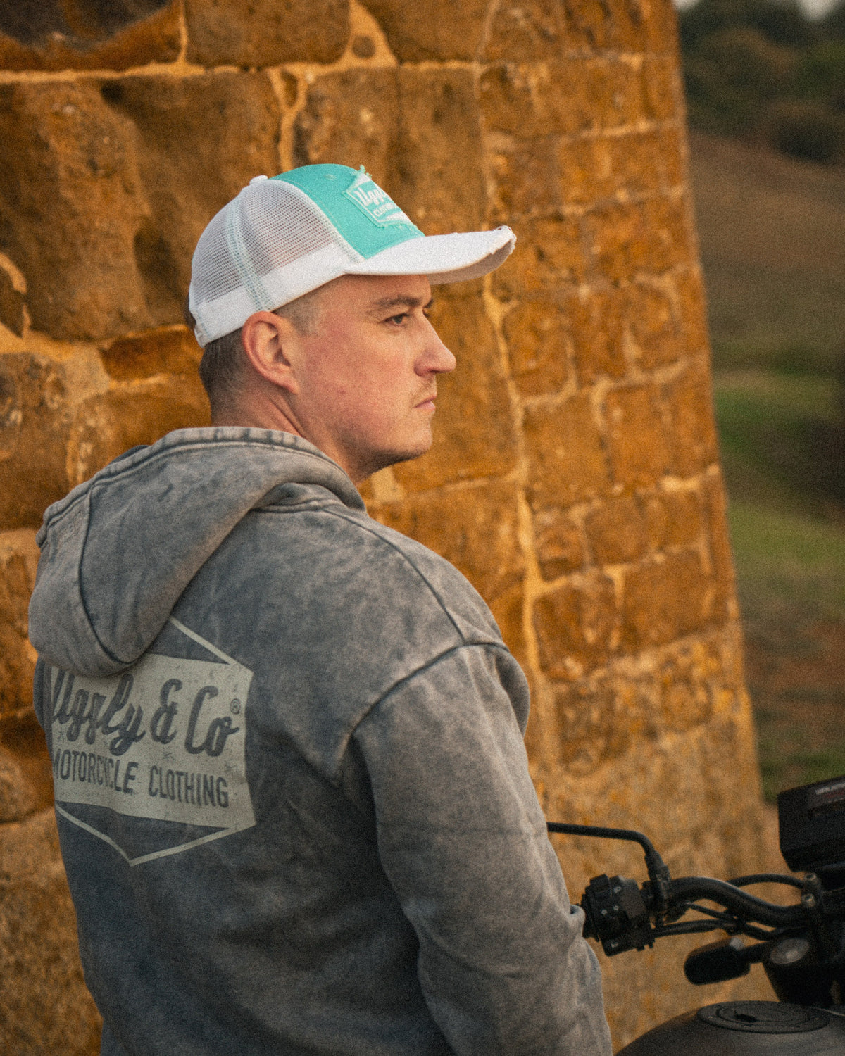 Biker wearing a vintage turquoise trucker cap next to a Royal Enfield Scram 411 scrambler motorcycle