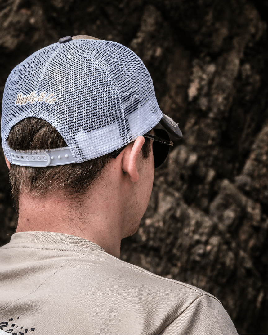 Rear view of a model wearing a beige and brown trucker cap with a diamond Uggly&Co logo and wave texture design, featuring a white mesh back and a curved peak, with a blurred rocky background.