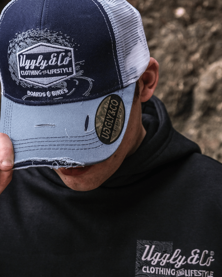 A model looking down while wearing a blue trucker hat with a white print of the Uggly&Co diamond logo and a light blue wave texture on the front, featuring a navy blue front panel, a sun-faded light blue peak, and a breathable white mesh back, with rocks in the background