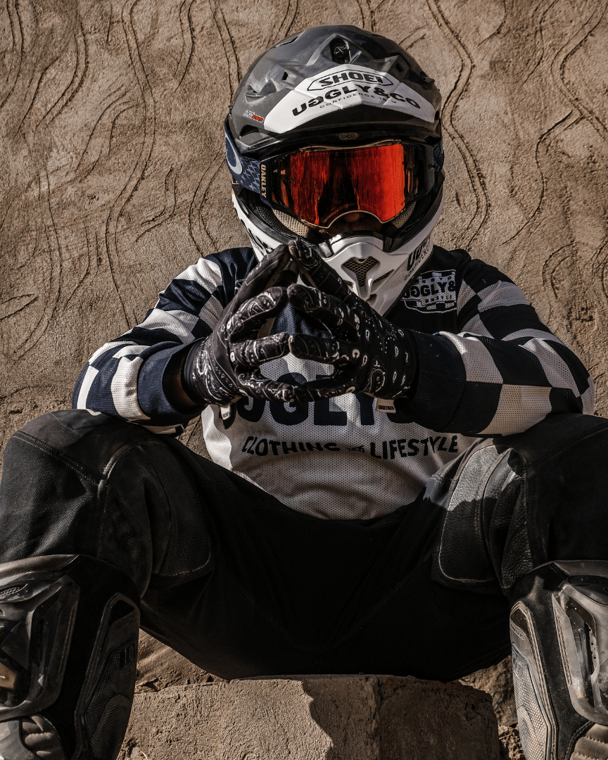 Motocross rider in full gear, helmet and goggles, sitting in the desert wearing a chequered Uggly&Co MX shirt