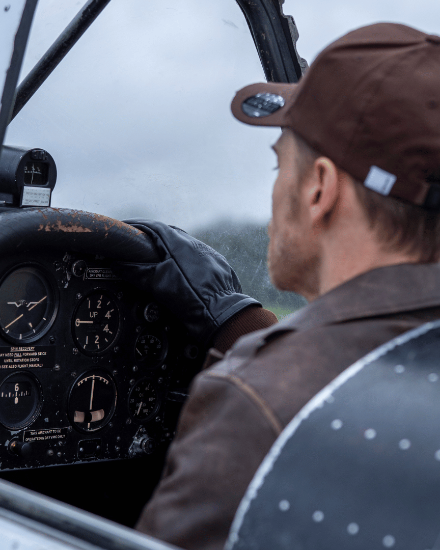 Rear view of a man wearing a brown 'Aviation' Diamond Trucker Hat with the Uggly&Co logo on the front and brown leather jacket sitting in the cockpit of a small airplane.